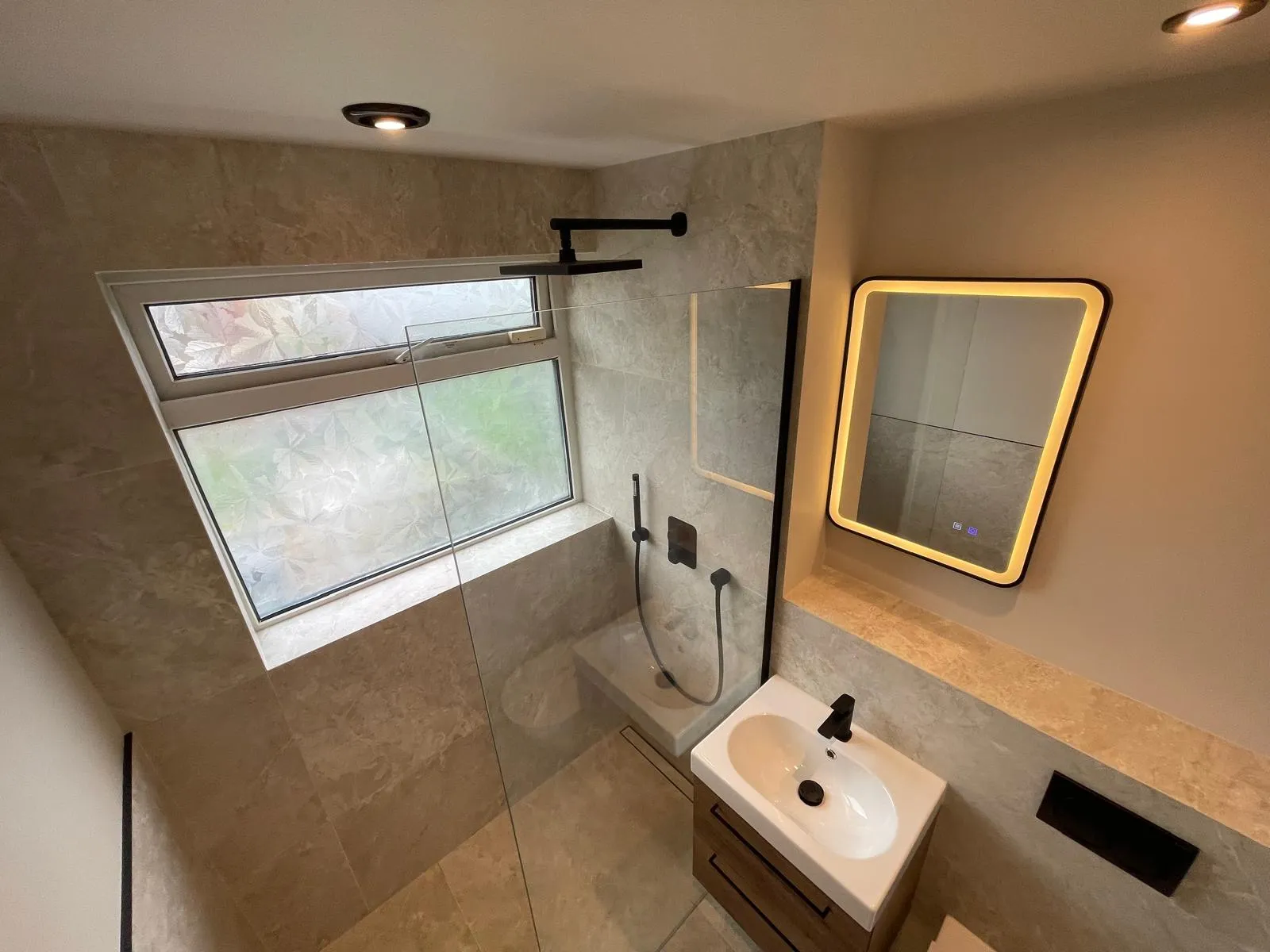 The same bathroom after the work, with stone-effect tiles, a walk-in shower and a lit mirror