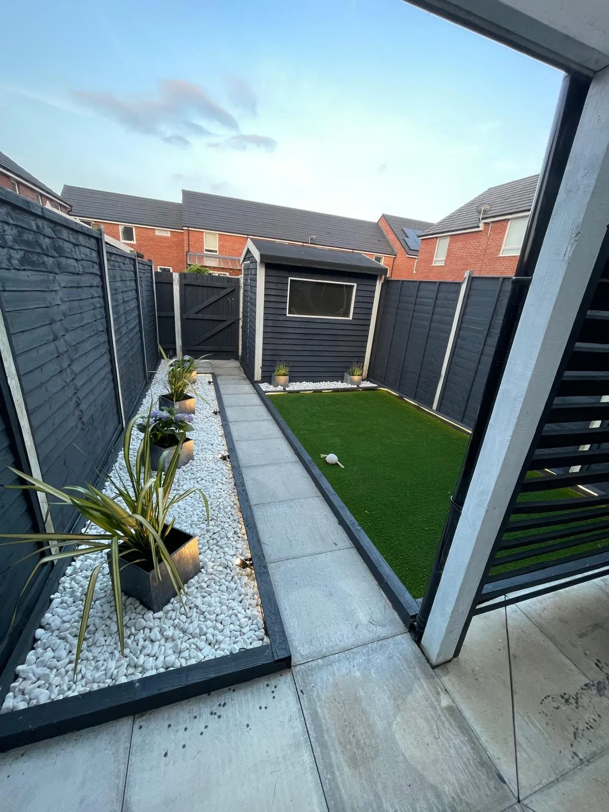 Porcelain patio with artificial lawn, white pebbles and a dark fence