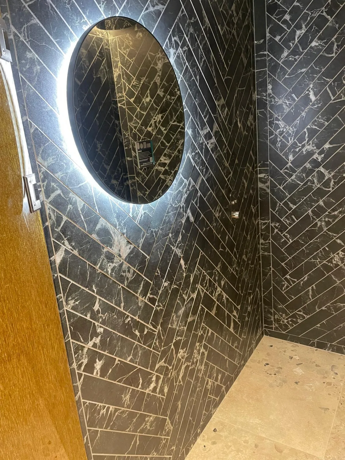 Shower tiled in black marble herringbone with a round lit mirror