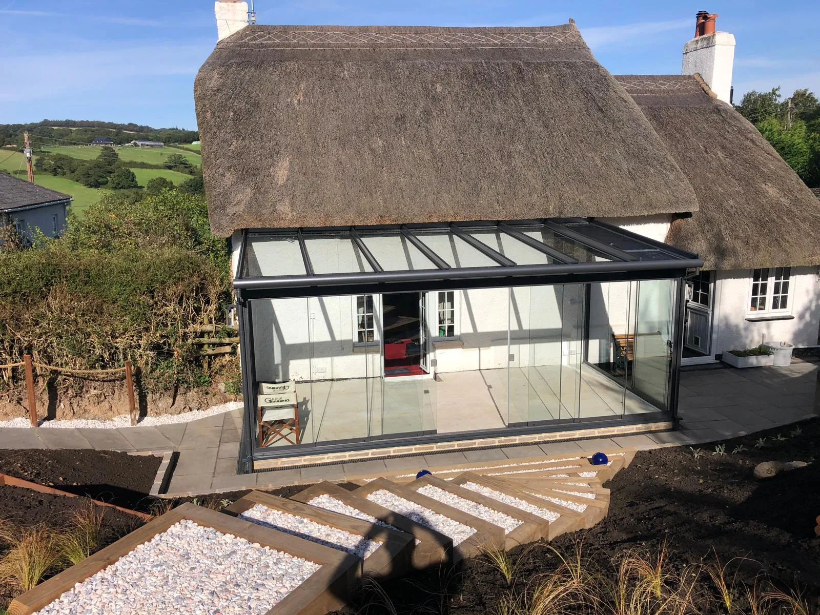 Frameless glass veranda built against a thatched cottage, with landscaped steps